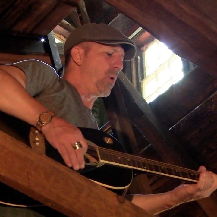 John playing a black acoustic guitar under the rafters of an attic, lit by a high window