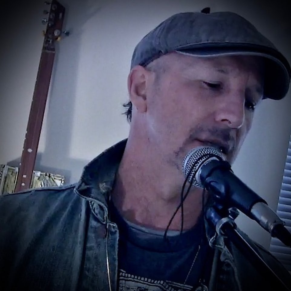 John Francis Mariano singing into a microphone in a flat cap, a cigar-box guitar on the wall behind him