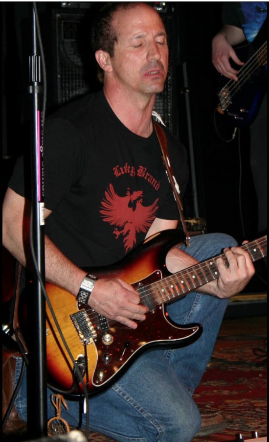 John playing a sunburst electric guitar on stage, eyes closed, singing