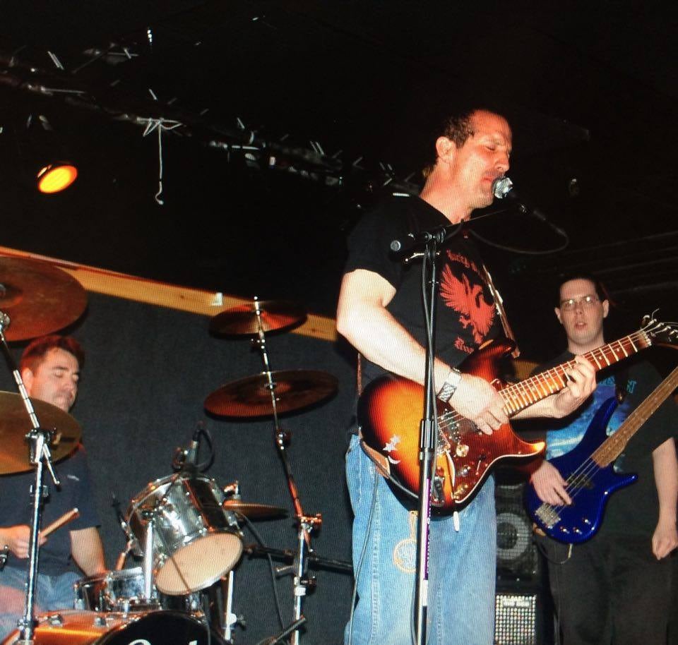 John singing and playing a sunburst electric guitar on stage, with the drummer and bassist behind him